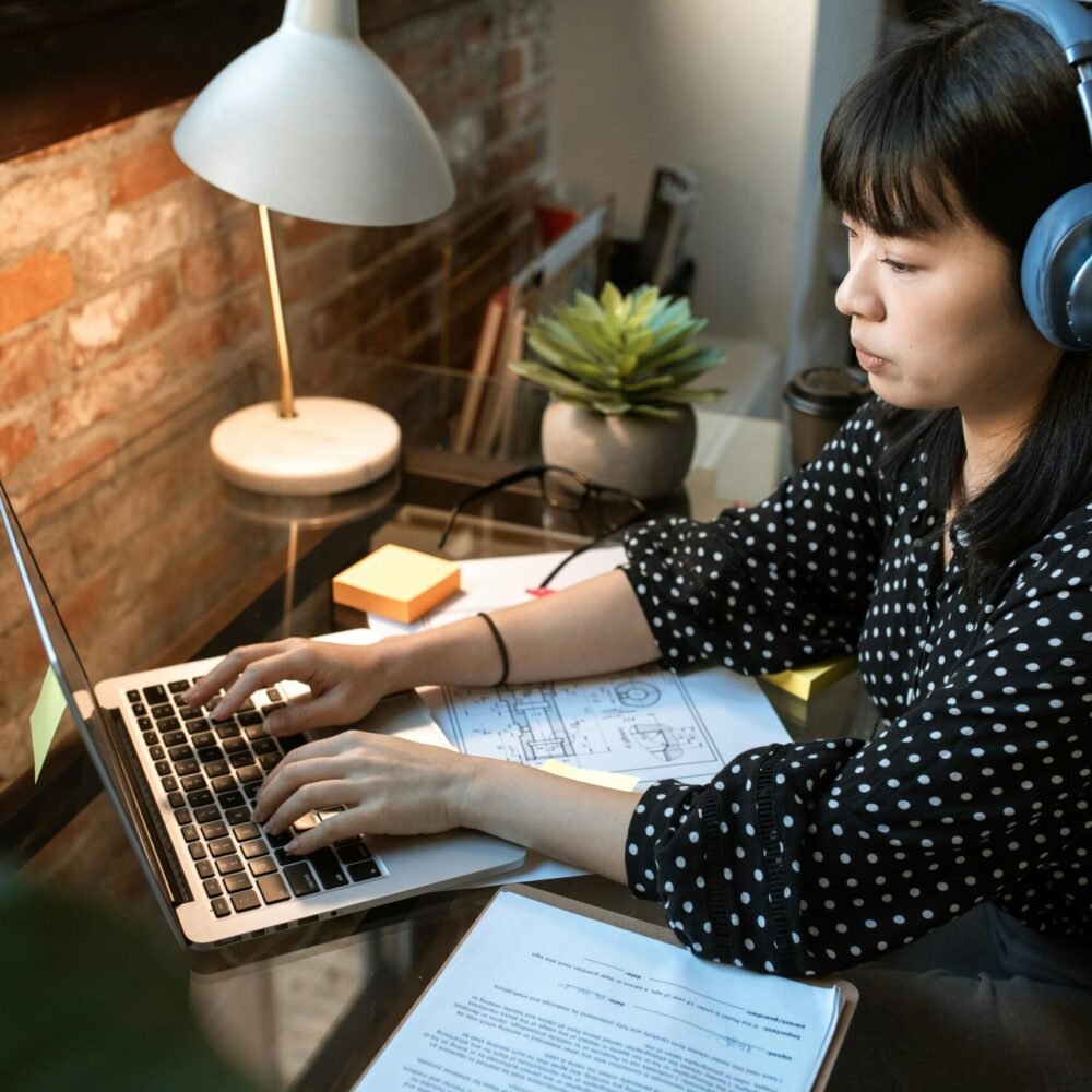 Focused woman wearing headphones, working on a laptop in a cozy home office setup with natural light.